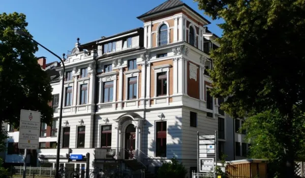 Historisches, mehrstöckiges Gebäude mit prachtvoller Fassade und Säulen am Eingang, vermutlich eine Einrichtung oder ein Zentrum. Umgeben von Bäumen und Straßenverkehr. Aufnahmewinkel bei Tageslicht mit blauem Himmel.