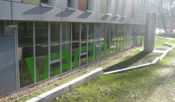 Moderne Gebäudefassade mit großflächigen Glasfronten und auffälligen grünen Einrichtungselementen im Inneren. Zeitgenössische Architektur mit Sonnenschutzelementen und gepflegter Außenanlage.