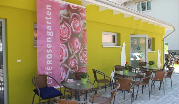 Grün gestrichene Terrasse eines Cafés mit dem Namen 'Rosengarten' auf einem rosa Banner. Gemütlicher Außenbereich mit mehreren Metallstühlen und kleinen Tischen für Gäste.