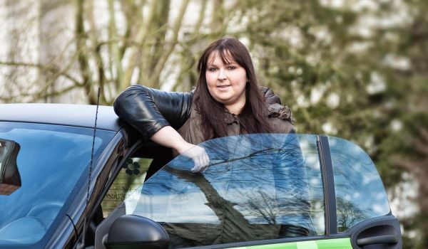 Eine junge Frau mit langen dunklen Haaren und einer schwarzen Lederjacke lehnt sich lächelnd an die offene Tür eines kleinen grünen Autos mit Blumenmotiven. Das Auto trägt eine Aufschrift, die auf Pflegedienst vacances hinweist. Im Hintergrund sind Bäume und eine leicht verschwommene städtische Umgebung zu sehen.
