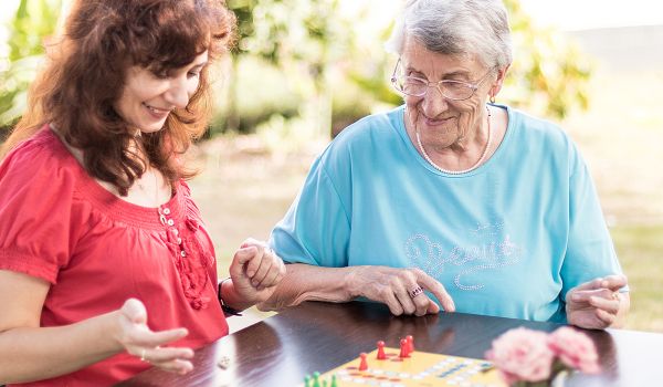 FsJ Freiwilligendienst in der Pflege in Bremen Zwei Personen sitzen an einem dunklen Holztisch und spielen ein Brettspiel mit bunten Spielfiguren. Im Vordergrund sind rosa Blumen zu sehen, im Hintergrund eine grüne Hecke.