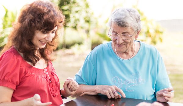 Zwei Personen sitzen an einem dunklen Holztisch und spielen ein Brettspiel mit bunten Spielfiguren. Im Vordergrund sind rosa Blumen zu sehen, im Hintergrund eine grüne Hecke.