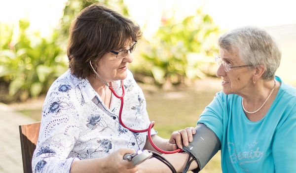 Eine ältere Frau in einem türkisfarbenen Shirt sitzt im Freien an einem Holztisch und lässt ihren Blutdruck messen. Eine Pflegekraft mit dunklen Haaren, Brille und einer weißen Bluse mit Blumenmuster nutzt ein Stethoskop und ein Blutdruckmessgerät, um die Werte zu überprüfen. Beide Frauen lächeln sich freundlich an. Im Hintergrund ist eine grüne Gartenlandschaft zu sehen.