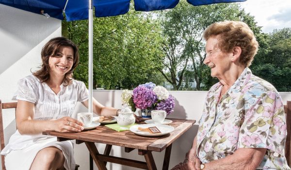 Zwei Frauen sitzen an einem Holztisch auf einem Balkon unter einem blauen Sonnenschirm. Sie genießen gemeinsam Kaffee und Kekse, während sie sich fröhlich unterhalten. Eine der Frauen trägt ein weißes Kleid, die andere eine gemusterte Bluse. Auf dem Tisch stehen Tassen, ein Teller mit Gebäck und ein Strauß bunter Hortensien.