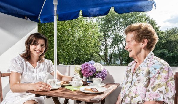 Zwei Frauen sitzen an einem Holztisch auf einem Balkon unter einem blauen Sonnenschirm. Sie genießen gemeinsam Kaffee und Kekse, während sie sich fröhlich unterhalten. Eine der Frauen trägt ein weißes Kleid, die andere eine gemusterte Bluse. Auf dem Tisch stehen Tassen, ein Teller mit Gebäck und ein Strauß bunter Hortensien.