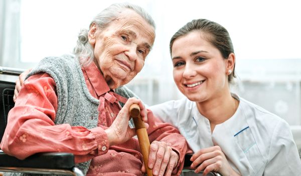 Eine ältere Frau mit grauen Haaren sitzt in einem Rollstuhl und hält einen Gehstock. Sie trägt eine rosa Bluse mit einer grauen Strickweste. Neben ihr kniet eine junge Pflegekraft in weißer Dienstkleidung, die freundlich lächelt. Beide wirken zufrieden und herzlich miteinander verbunden.