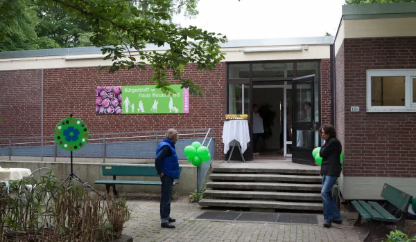 Eingang des 'Rosengartens' Gemeinschaftszentrums in einem eingeschossigen Backsteingebäude mit buntem Schild. Besucherinnen und Besucher stehen vor der Eingangstreppe mit grünen Dekorationselementen. Einladende Atmosphäre einer sozialen Einrichtung in grüner Umgebung.