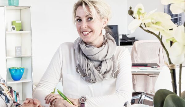Eine freundliche Frau mit blonden Haaren, die zu einem lockeren Dutt gebunden sind, sitzt in einem modernen Büro. Sie trägt eine weiße Bluse, einen grauen Schal und Perlenohrringe. In ihrer Hand hält sie einen grünen Stift, während sie mit einer anderen Person spricht. Im Hintergrund sind ein Schreibtisch mit Laptop, ein Regal mit Dekoartikeln und eine weiße Orchidee zu sehen. Das Büro ist hell und stilvoll eingerichtet.