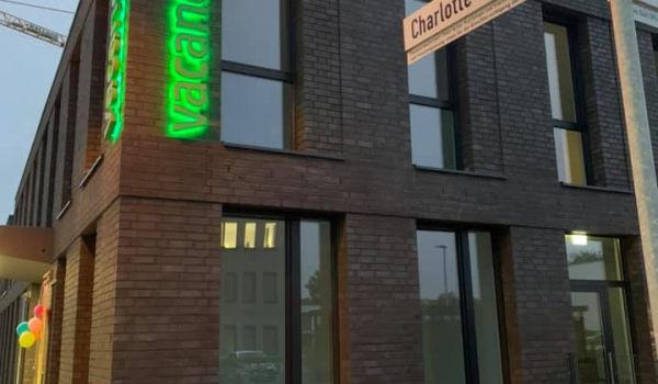 Modernes zweigeschossiges Backsteingebäude Tagespflege Bremen Lesum' mit beleuchtetem grünen vacances-Logo bei Dämmerung. Zeitgemäße Architektur mit großen Fenstern und gepflastertem Vorplatz.