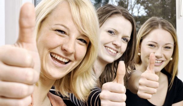 Drei junge Frauen mit hellen Haaren lächeln fröhlich und zeigen den Daumen nach oben.