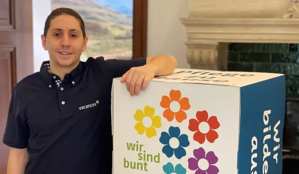Ein junger Mann mit dunklen Haaren und einem dunkelblauen Poloshirt mit ‘vacances’-Logo lehnt an einem großen, weißen Würfel. Der Würfel ist mit bunten Blumen und den Schriftzügen ‘wir sind bunt’ und ‘wir bilden aus’ bedruckt. Er steht in einem Innenraum mit einem Kamin im Hintergrund und lächelt freundlich in die Kamera.