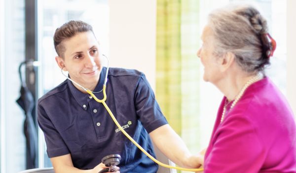 Person mit dunklem Haar und blauem Poloshirt mit Stethoskop die den Blutdruck einer älteren Person misst mit rosa Oberteil und Perlenkette.