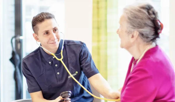 Person mit dunklem Haar und blauem Poloshirt mit Stethoskop die den Blutdruck einer älteren Person misst mit rosa Oberteil und Perlenkette.