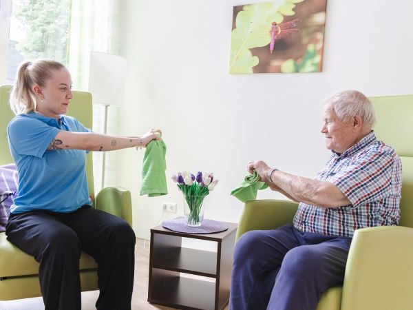 Vital Coach in blauer Arbeitskleidung führt mit einem älteren Mann im Sessel eine Bewegungsübung mit grünen Handtüchern durch.
