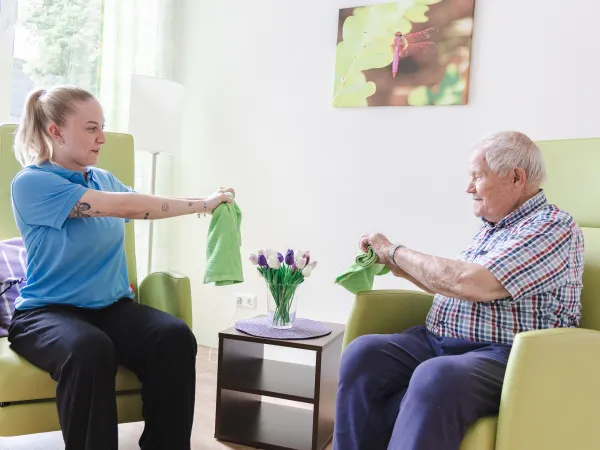 Vital Coach in blauer Arbeitskleidung führt mit einem älteren Mann im Sessel eine Bewegungsübung mit grünen Handtüchern durch.