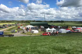 Veranstaltungsgelände mit Zelten und Fahrzeugen auf ländlicher Wiese unter dramatischem Wolkenhimmel.