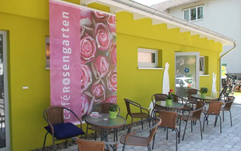 Grün gestrichene Terrasse eines Cafés mit dem Namen 'Rosengarten' auf einem rosa Banner. Gemütlicher Außenbereich mit mehreren Metallstühlen und kleinen Tischen für Gäste.