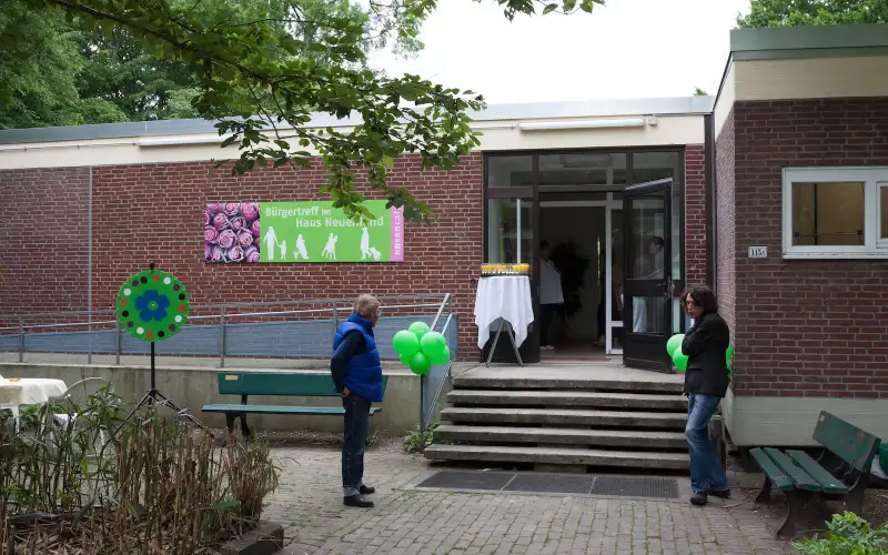 Eingang des 'Rosengartens' Gemeinschaftszentrums in einem eingeschossigen Backsteingebäude mit buntem Schild. Besucherinnen und Besucher stehen vor der Eingangstreppe mit grünen Dekorationselementen. Einladende Atmosphäre einer sozialen Einrichtung in grüner Umgebung.