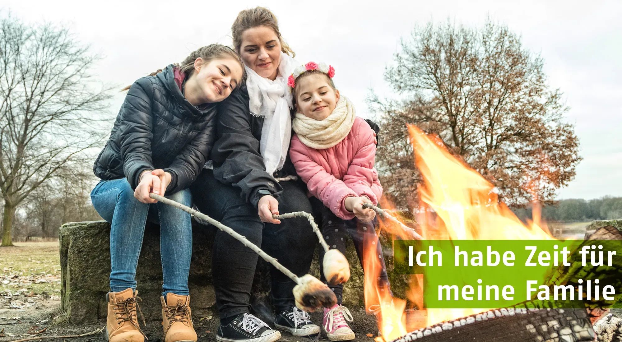 Eine Mutter sitzt mit ihren zwei Töchtern im Freien am Lagerfeuer und röstet Stockbrot. Alle drei wirken glücklich und entspannt. Der Text im Bild lautet: ‚Ich habe Zeit für meine Familie‘. Die Szene vermittelt Wärme, Zusammenhalt und Familienzeit in der Natur.