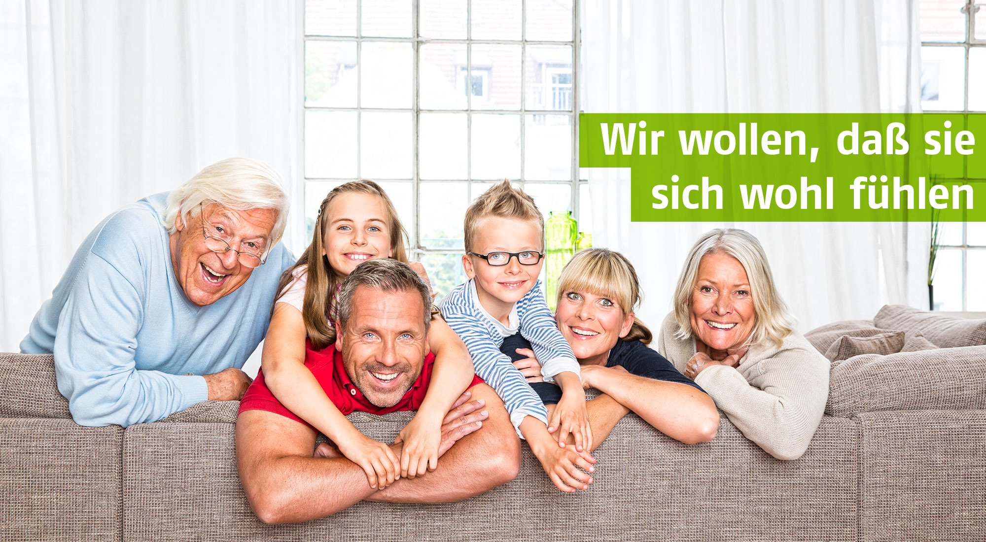 Glückliche Mehrgenerationen-Familie auf einem Sofa - Großeltern, Eltern und Kinder lächeln gemeinsam in einer hellen Wohnzimmeratmosphäre. Mit grünem Textbalken: 'Wir wollen, daß sie sich wohl fühlen'