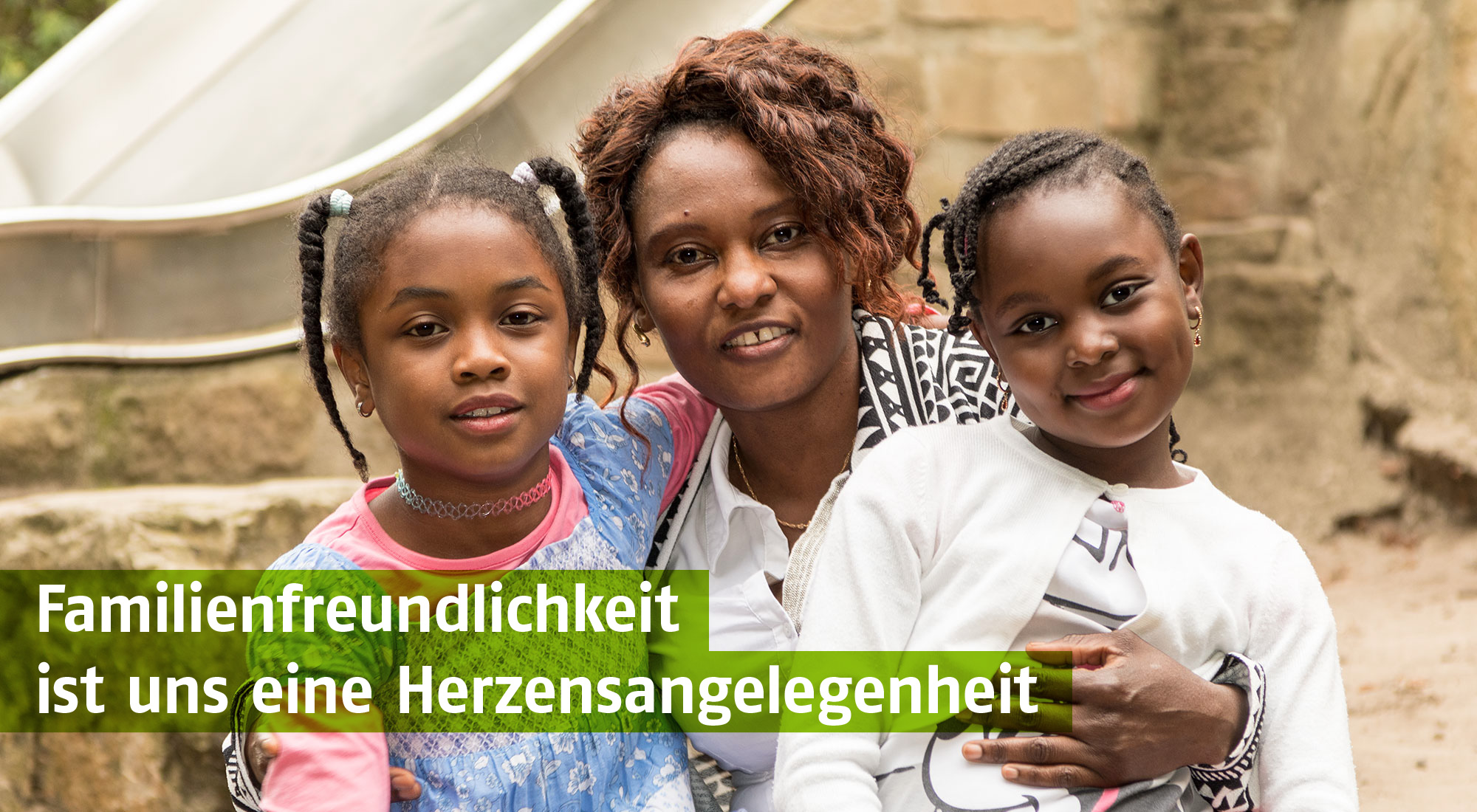 Lächelnde Mutter mit zwei Töchtern in einer Freizeitumgebung mit Spielplatz. Die Familie strahlt Zusammenhalt und Freude aus. Grüner Textbalken: 'Familienfreundlichkeit ist uns eine Herzensangelegenheit'