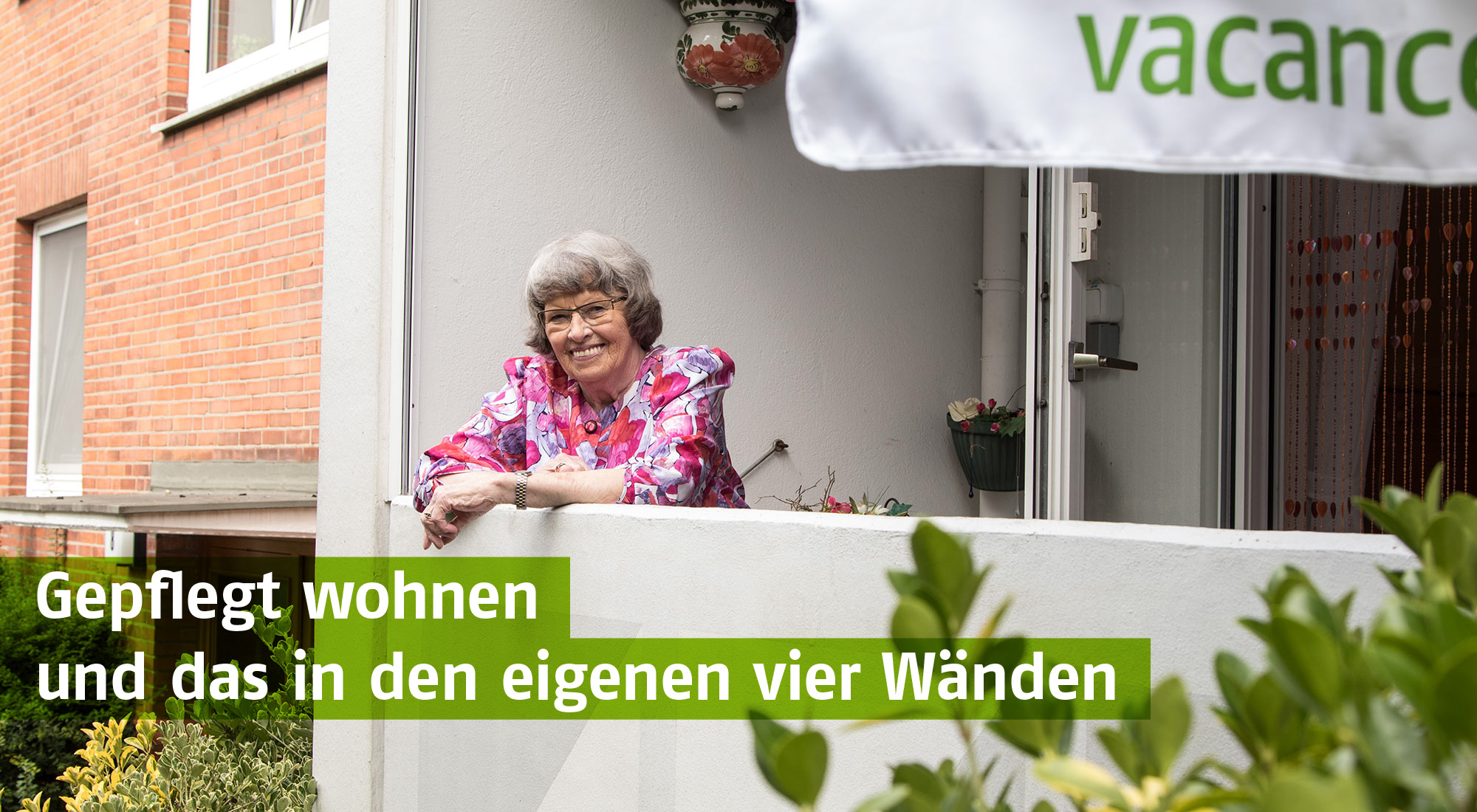 Lächelnde Seniorin mit Brille auf ihrem Balkon, gekleidet in einem farbenfrohen rosa Oberteil. Die Dame genießt sichtlich ihr Zuhause. Grüner Textbalken: 'Gepflegt wohnen und das in den eigenen vier Wänden'