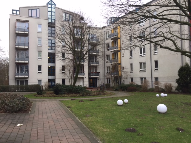 Moderne Wohnanlage mit mehreren mehrstöckigen Gebäuden in hellen Farbtönen. Gepflegte Außenanlage mit Rasenfläche, dekorativen weißen Kugeln und gepflasterten Wegen zwischen den Häusern.