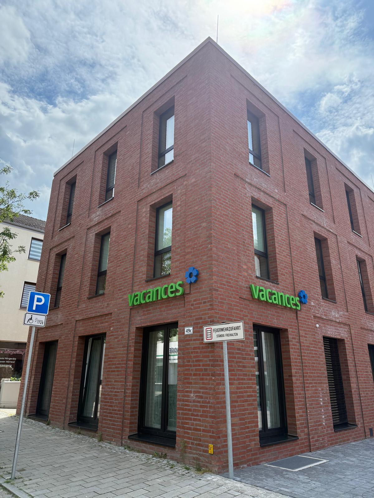 Modernes zweigeschossiges Backsteingebäude Tagespflege Bremen Vegesack mit grünen vacances-Logo. Zeitgemäße Architektur mit großen Fenstern und gepflastertem Vorplatz.