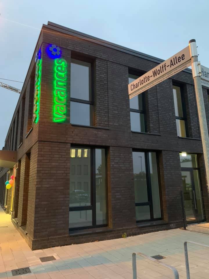 Tagespflege Lesum Modernes zweigeschossiges Backsteingebäude Tagespflege Bremen Lesum' mit beleuchtetem grünen vacances-Logo bei Dämmerung. Zeitgemäße Architektur mit großen Fenstern und gepflastertem Vorplatz.