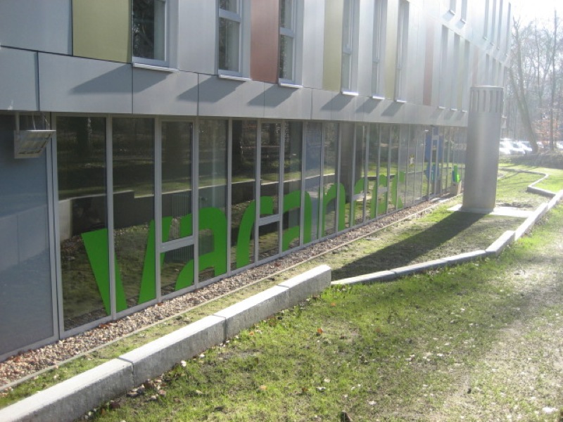 Vacances Büro Blumenthal Moderne Gebäudefassade mit großflächigen Glasfronten und auffälligen grünen Einrichtungselementen im Inneren. Zeitgenössische Architektur mit Sonnenschutzelementen und gepflegter Außenanlage.