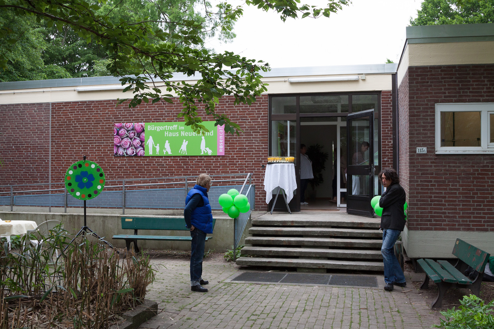 Eingang des 'Rosengartens' Gemeinschaftszentrums in einem eingeschossigen Backsteingebäude mit buntem Schild. Besucherinnen und Besucher stehen vor der Eingangstreppe mit grünen Dekorationselementen. Einladende Atmosphäre einer sozialen Einrichtung in grüner Umgebung.
