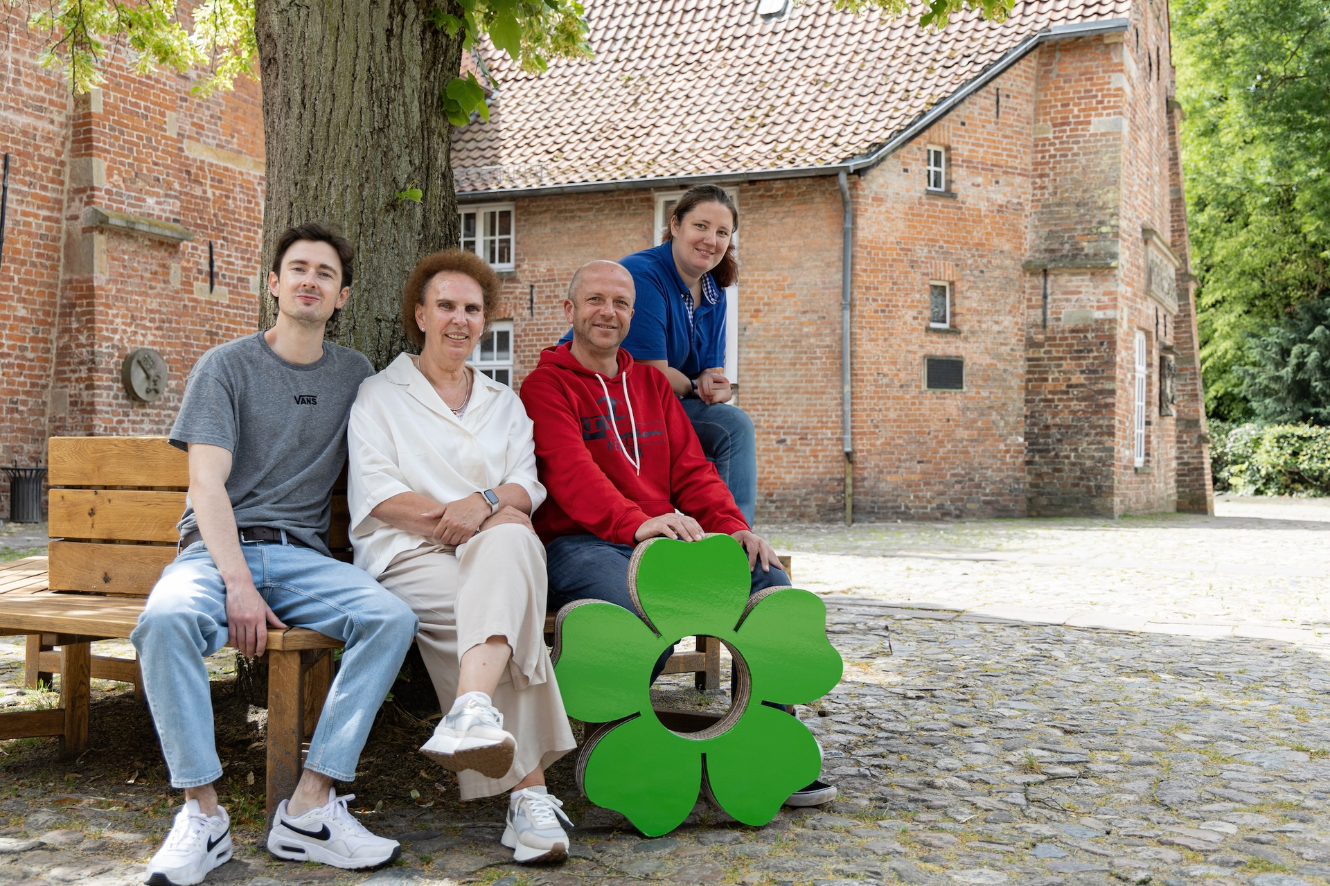 Vier Teammitglieder des Pflegedienstes sitzen auf einer Holzbank vor einem historischen Backsteingebäude. Die Gruppe präsentiert stolz das die grüne Blume von vacances.