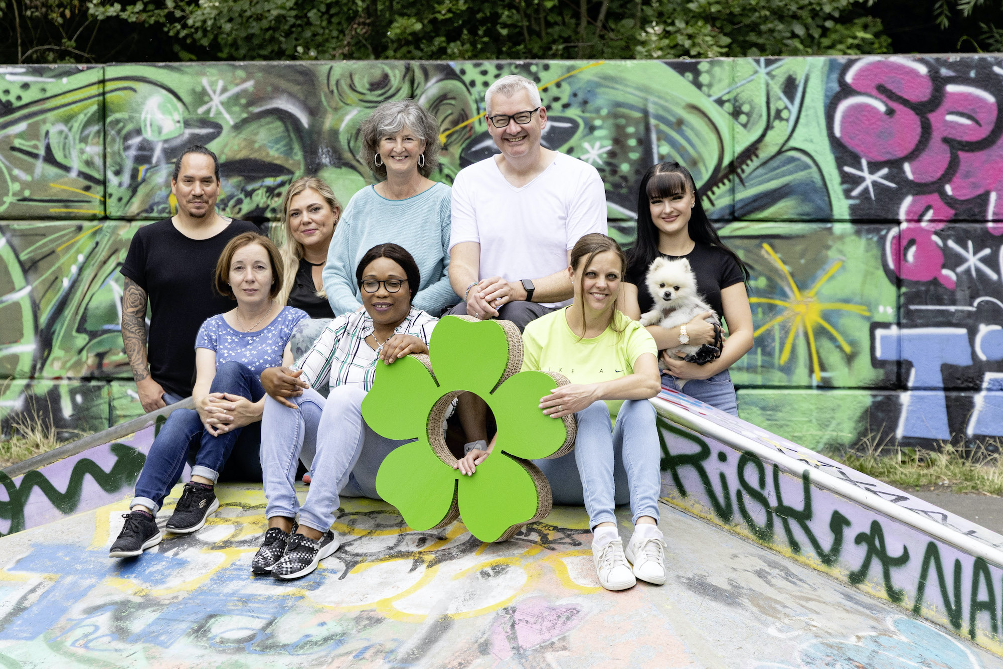 Diverse Gruppe posiert in Skatepark vor Graffiti-Wand, mit grüner Blumenform und kleinem weißen Hund. Team Huchting