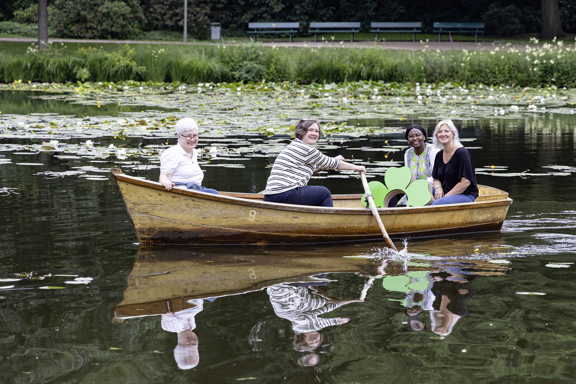 Team-Schwachhausen Vier Frauen im Holzboot auf Teich mit Seerosen, grünes Blumen-Logo sichtbar, Parkbänke im Hintergrund.